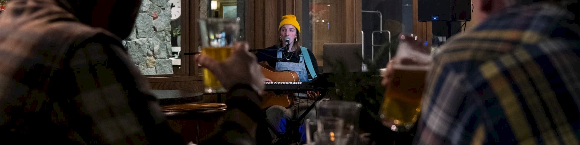 A singer in a yellow beanie performs with an acoustic guitar onstage in a lounge, watched by diners.