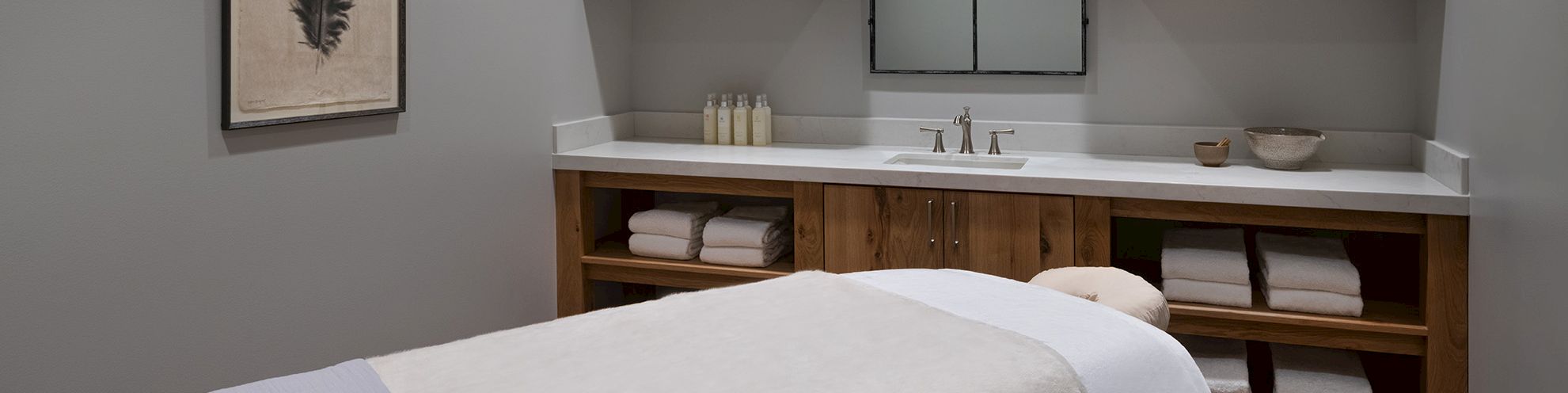 A modern spa bathroom with a wooden vanity, white countertop, twin sinks, stacked towels, and framed wall art.
