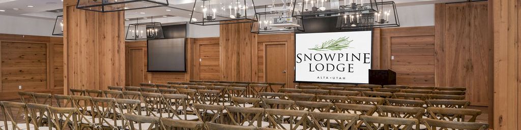 An empty lodge banquet hall with rows of chairs and hanging lantern-style lights, featuring a Snowpine Lodge logo backdrop.