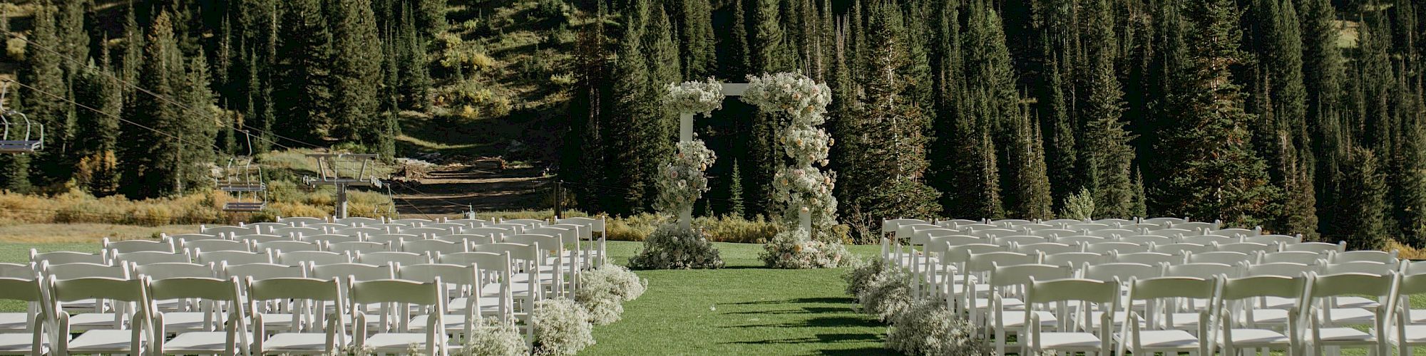 Outdoor wedding setup with white chairs, a floral arch, green lawn, and pine-covered mountain backdrop under a clear sky.