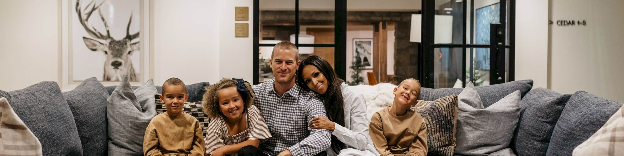 A family of five poses on a cozy gray sofa in a bright living room with modern glass doors behind them.