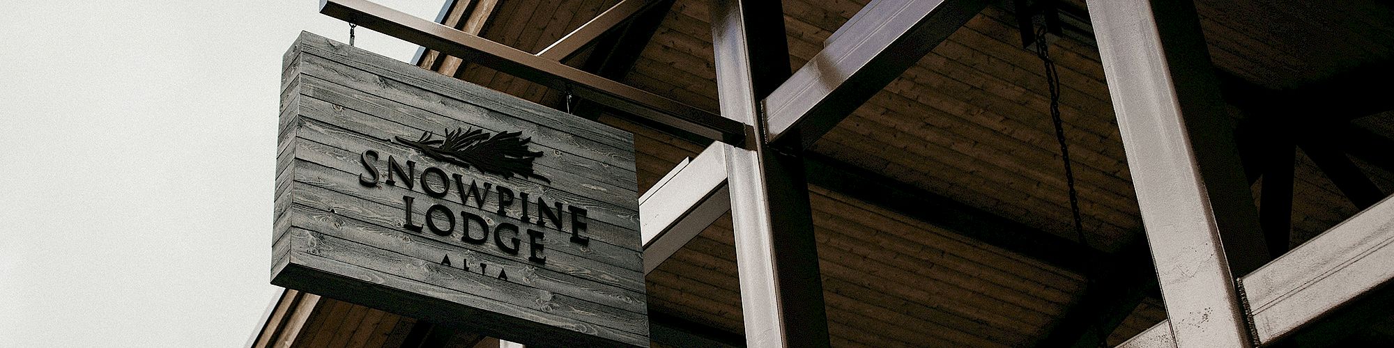A wooden sign reading &ldquo;Snowpine Lodge&rdquo; hangs above steel beams and a chain near an industrial building entrance.