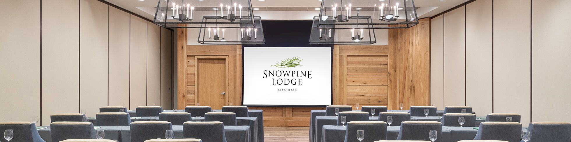 Modern meeting room with gray chairs, hanging chandeliers, and a screen reading &ldquo;Snowpine Lodge&rdquo; at the front.