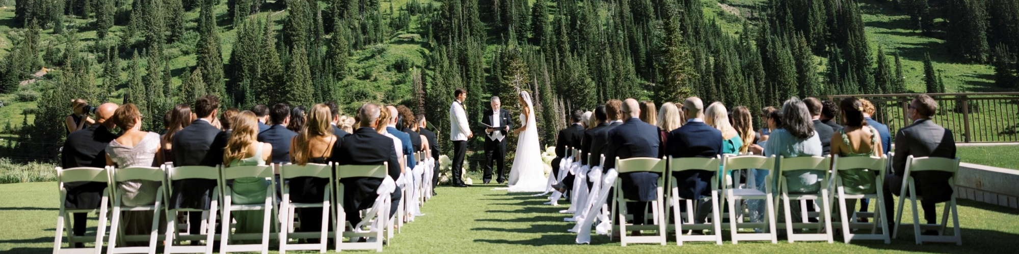 An outdoor wedding ceremony with the couple standing at an aisle, surrounded by guests, on a sunny mountain hillside.