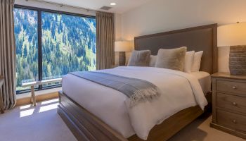 A modern bedroom with a large bed and lamps, plus a window with curtains showing pine-covered mountains outside.