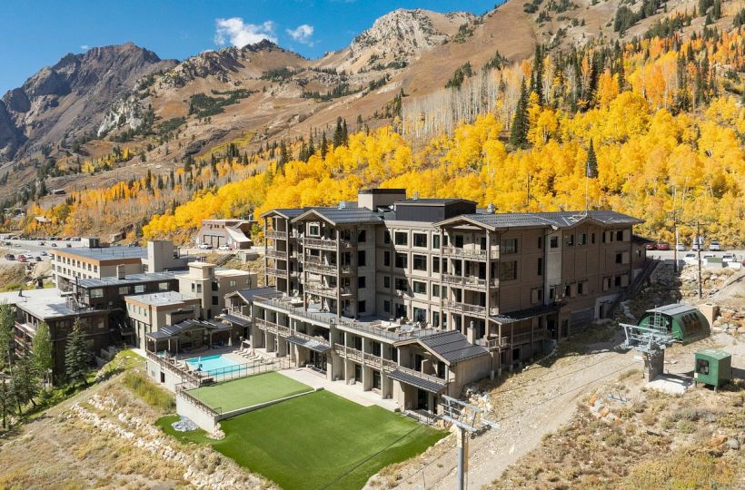 Mountain resort with balconies and a tennis court, surrounded by autumn-yellow trees and steep peaks under a blue sky.