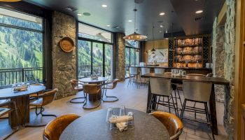 Cozy mountain lodge bar and dining area with stone walls, leather stools, and big windows showing green forest views.