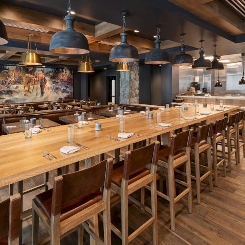 A rustic restaurant dining room with long wooden tables, chairs, table settings, and many hanging metal pendant lights.