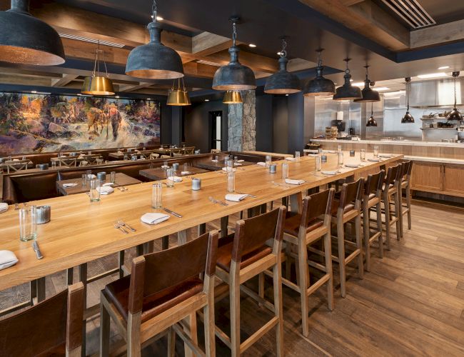 A rustic restaurant dining room with long wooden tables, chairs, table settings, and many hanging metal pendant lights.