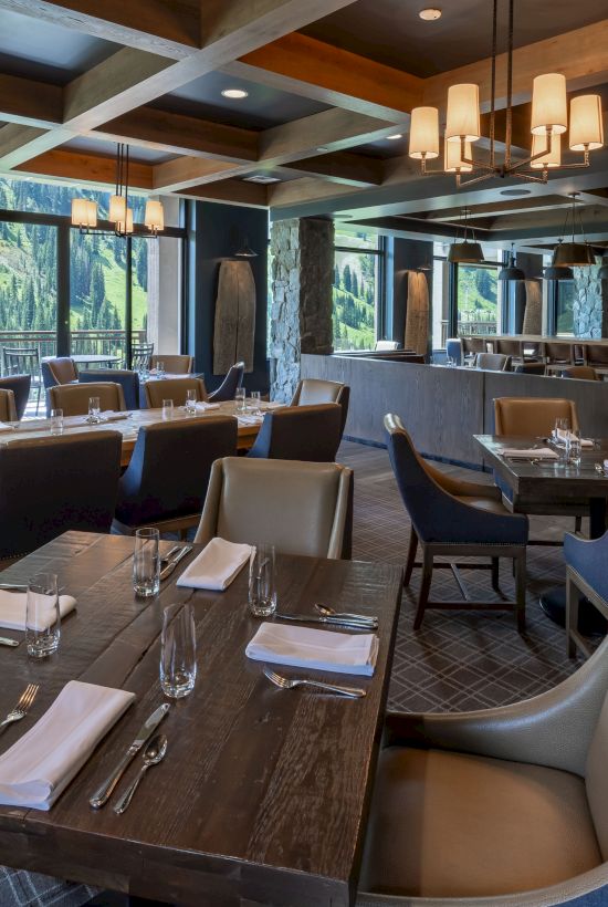A modern alpine restaurant dining area with wooden tables, upholstered chairs, warm pendant lighting, and large windows showing forested mountains.