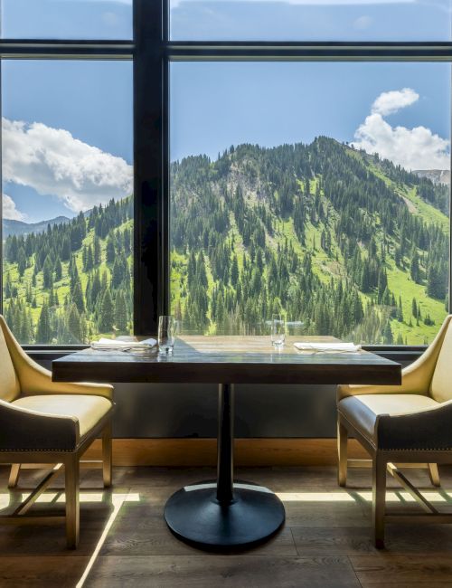 A sunlit restaurant table with two chairs sits by a big window, facing lush green mountains and clouds outside.