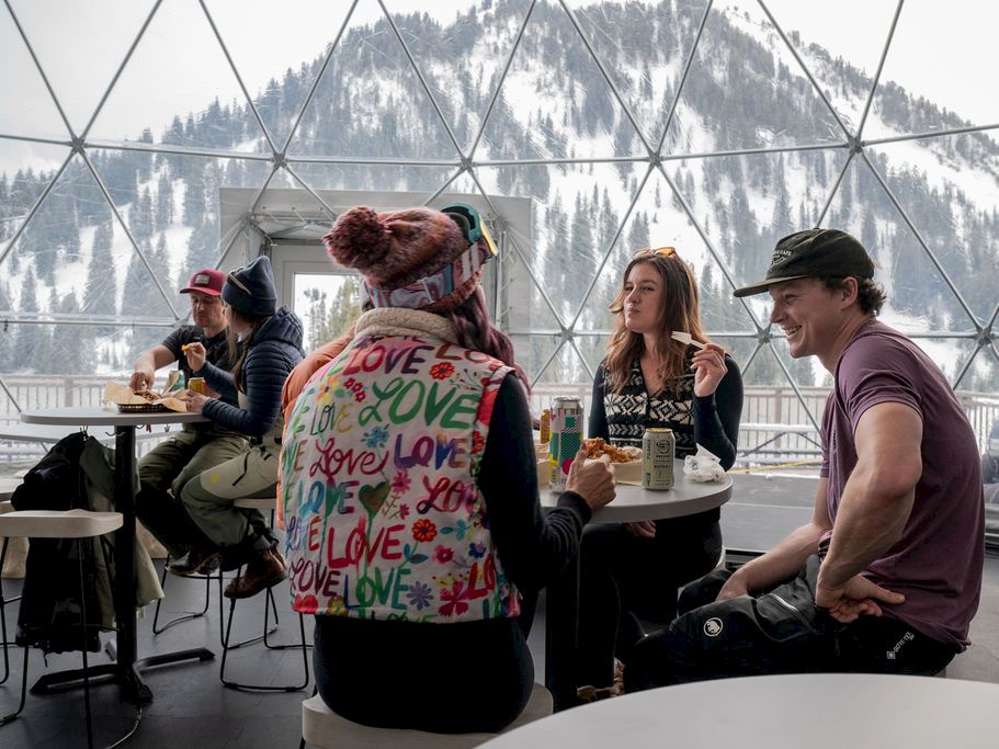 People eat and drink at a mountain caf&eacute; inside a glass dome with snowy peaks outside, chatting and smiling.