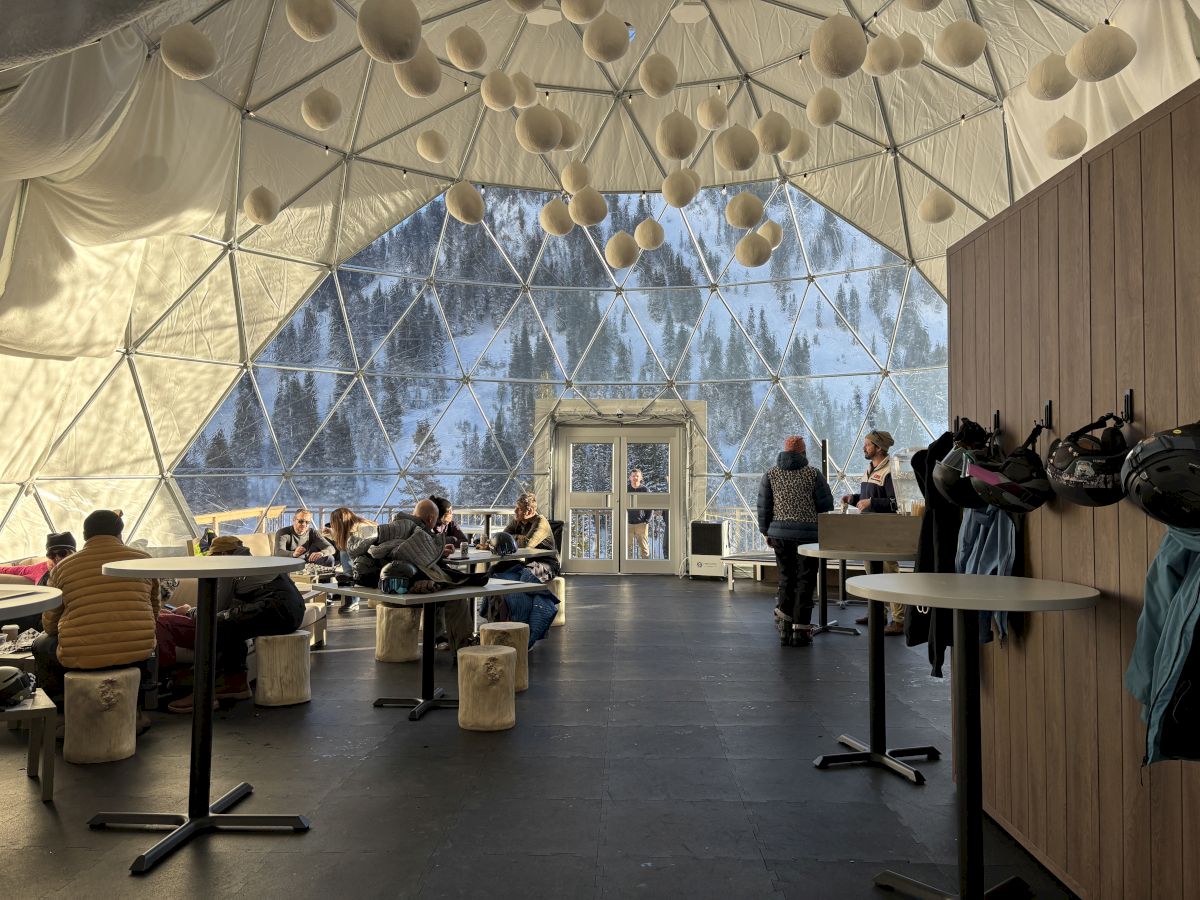 Inside a geodesic dome lounge, people eat while ski helmets hang nearby and a snowy forest is visible through the windows.
