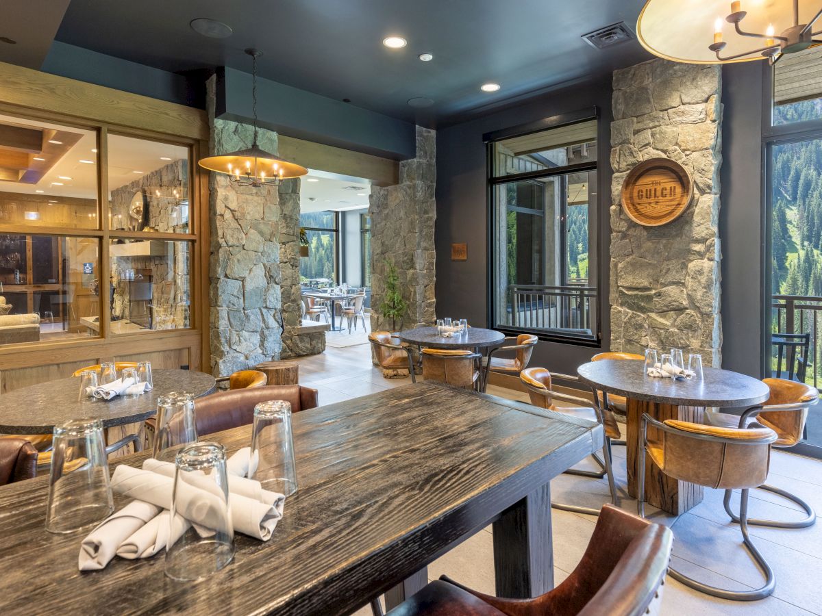 A cozy lodge dining room with wooden tables, stone walls, hanging lamps, and large windows overlooking pine-covered mountains.