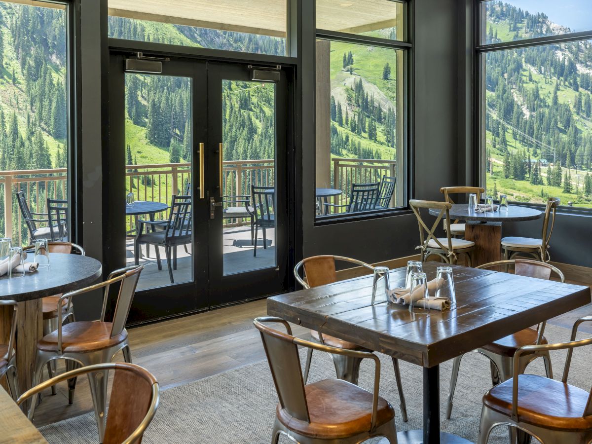 A cozy restaurant lounge with wooden tables and chairs, floor-to-ceiling doors/windows showing green alpine mountains outside.