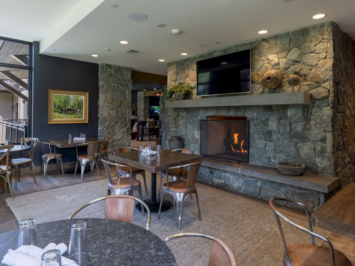 A cozy lounge restaurant with stone walls, a lit fireplace, large TV, and tables with chairs arranged for dining.