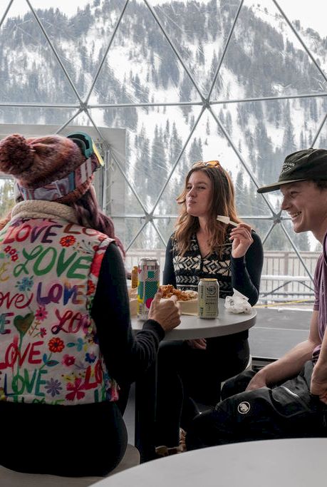 People dine and chat inside a glass geodesic tent with mountain views, sharing drinks and snacks.
