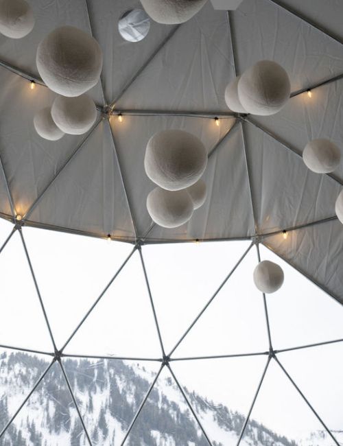 A lit dome interior with hanging paper lantern-like pom-poms and string lights, revealing snow-covered mountains through the windows.