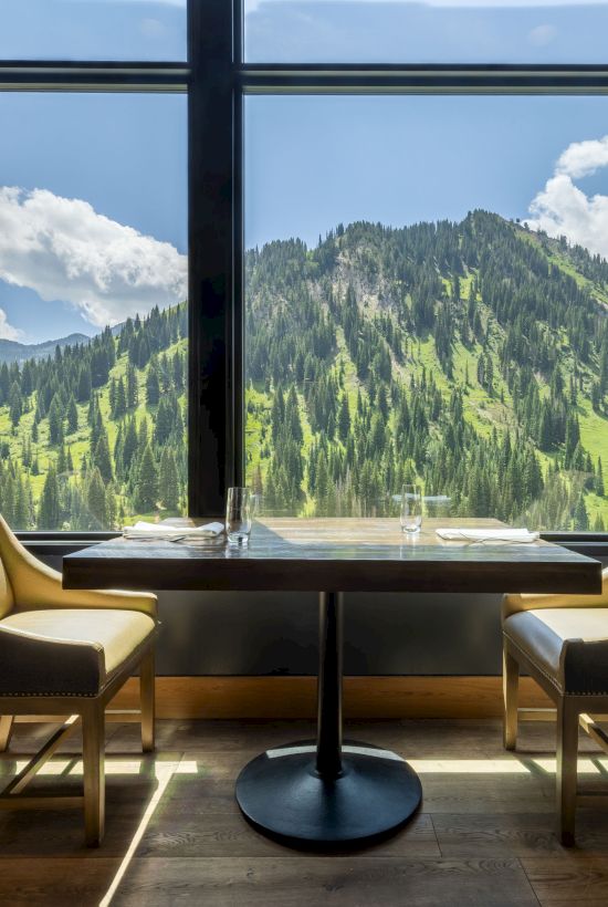 A cozy restaurant dining area with two chairs and a table by large windows, overlooking a lush green mountain valley under blue skies.