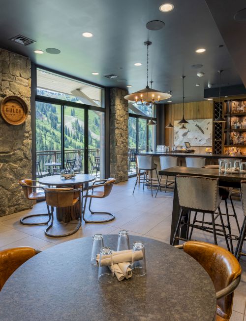 A cozy lodge-style bar interior with stone pillars, mountain-view windows, hanging lights, and stocked liquor shelves, with tables and stools.