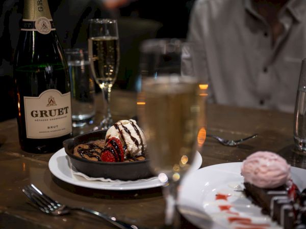 A champagne bottle and flute glasses sit by a plated chocolate dessert topped with berries, with a water glass in the background.