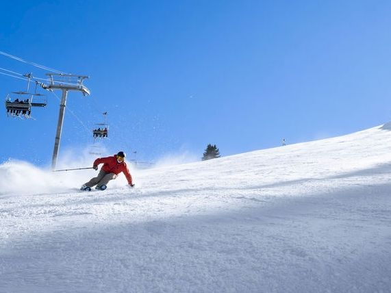 A skier in a red jacket carves fresh snow on a sunny slope with ski lifts overhead.
