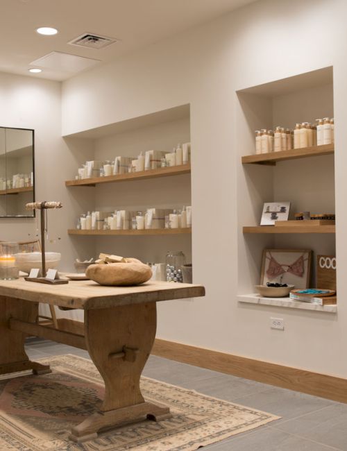 A bright spa shop with shelves of skincare bottles, a candle-lit display, and a wooden central table with a rug.