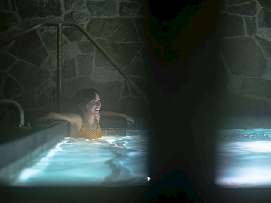 A woman in a yellow swimsuit sits in an indoor pool, smiling as light filters through the water and steam.