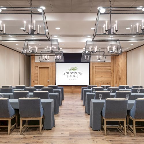 Modern banquet hall with gray chairs and hanging chandeliers, with a screen reading &ldquo;Snowpine Lodge.&rdquo;