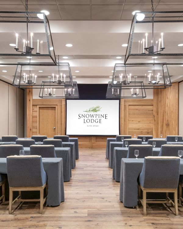 Modern banquet hall with gray chairs and hanging chandeliers, with a screen reading &ldquo;Snowpine Lodge.&rdquo;