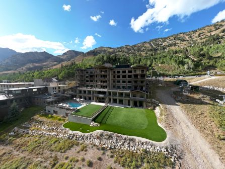 A mountain resort with apartment buildings, a small manicured grass golf green, cars, and a backdrop of green hills under blue sky.