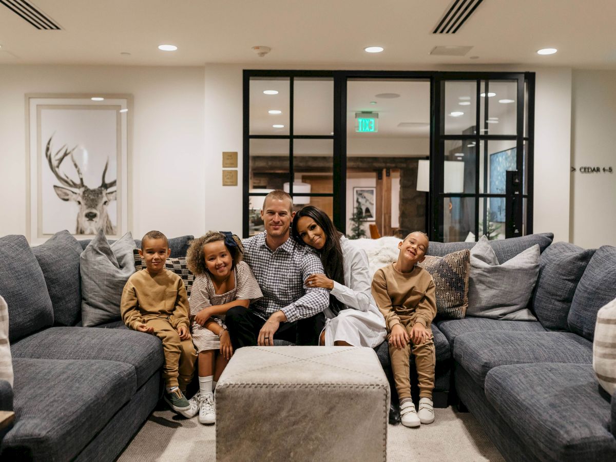 A family of five smiles on a gray couch in a bright lobby while the man and woman pose with the kids.