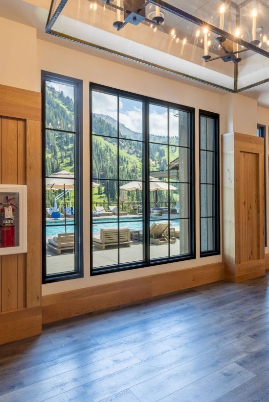 An indoor lodge hallway with wood panels, big windows showing a pool terrace, a fire extinguisher, and hanging lights above.