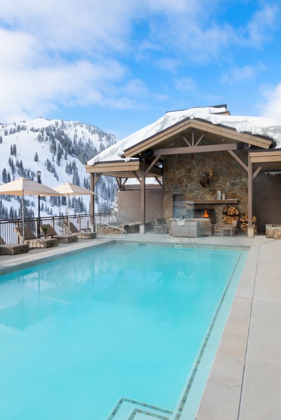 Snowy mountain resort patio with a heated pool, lounge chairs under umbrellas, and a stone fireplace by the slopes.