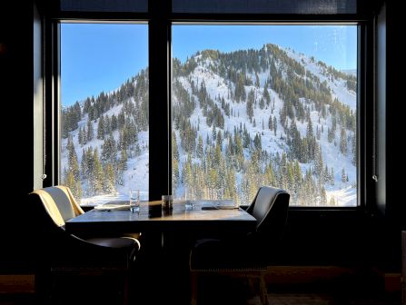 A restaurant table with glasses sits by large windows, overlooking a snowy alpine mountainside with pine trees and bright sky.