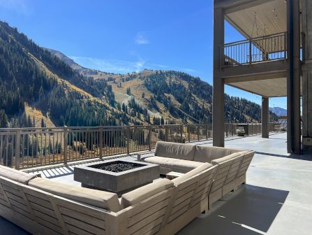 A sunny mountain valley view from a modern outdoor terrace with a lounge sofa, fire pit, and railing.