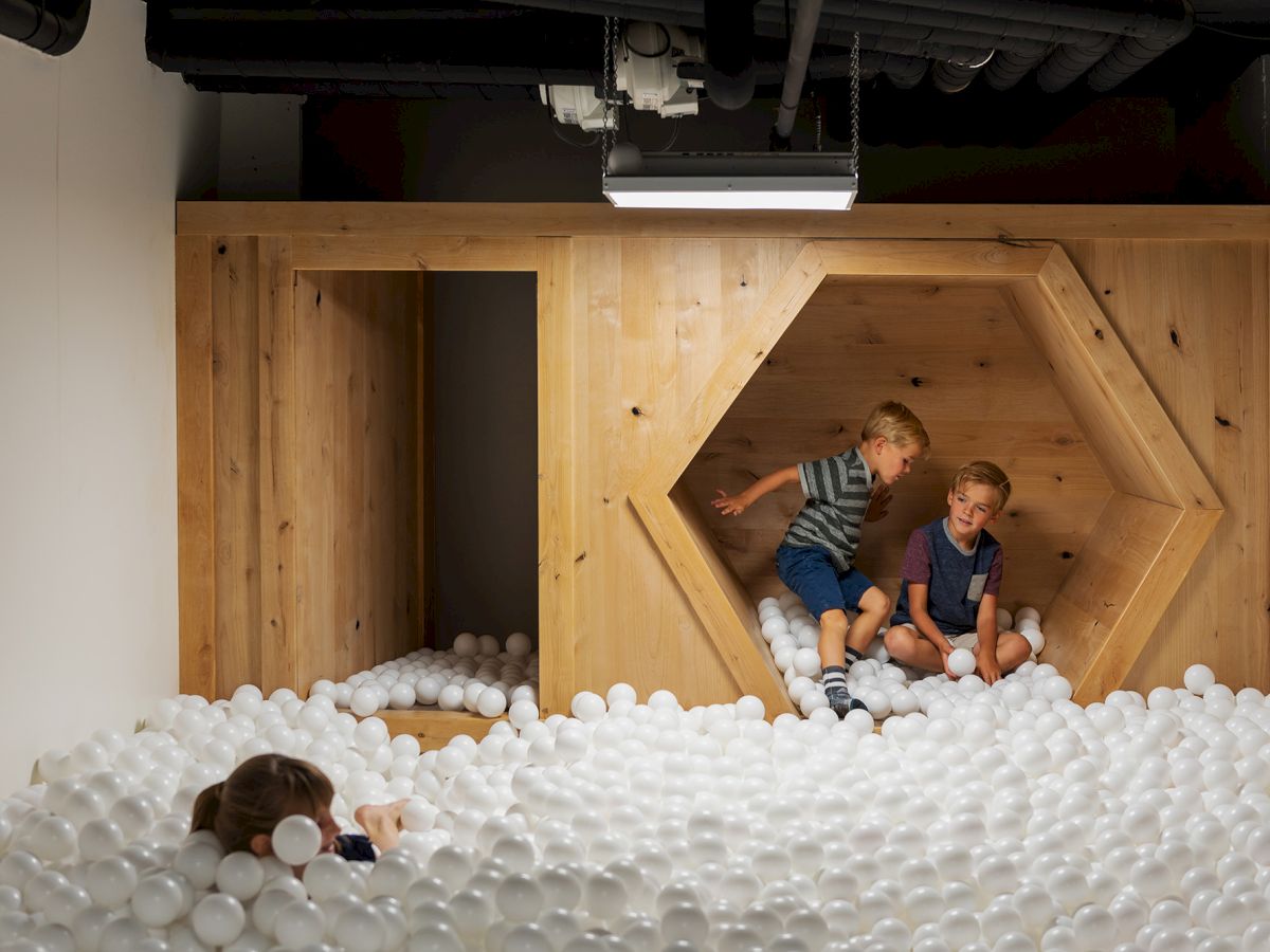 Two kids play in a white ball pit inside a wooden play structure.