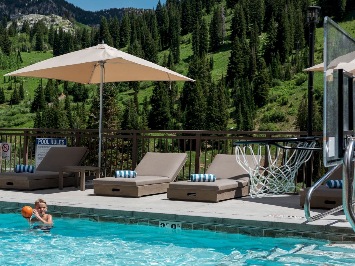 Outdoor pool with beige loungers under a shade umbrella, plus alpine forest mountains in the background.