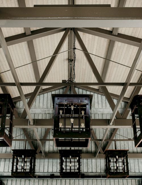Steel trusses and hanging metal frames holding rows of bulb-like lamps sway under chains in a factory hall.