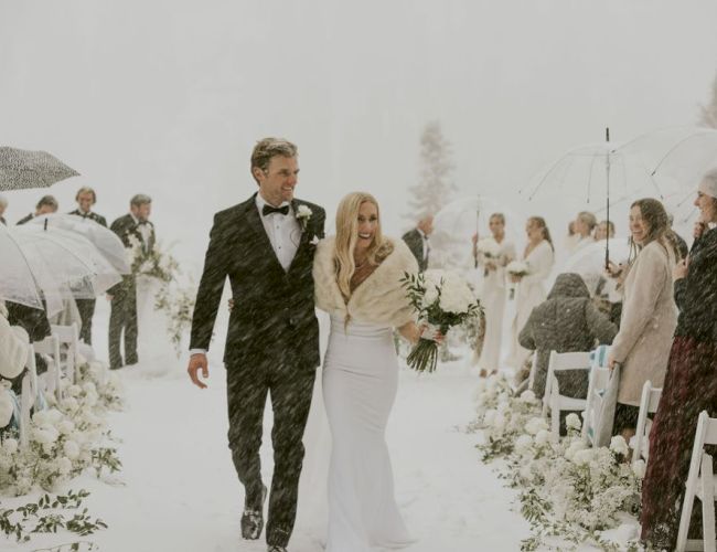 A snowy outdoor wedding procession in white: a groom in a tuxedo and a bride holding flowers walk down the aisle.