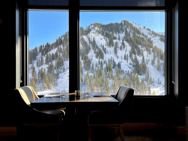 A restaurant table with glasses sits by large windows, overlooking a snowy alpine mountainside with pine trees and bright sky.