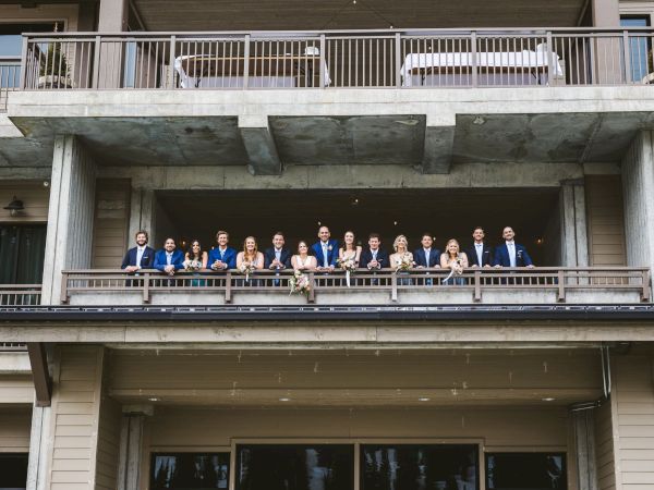 A wedding party of about 14 people in blue suits and formal dresses, holding bouquets, poses on a building&rsquo;s balcony.