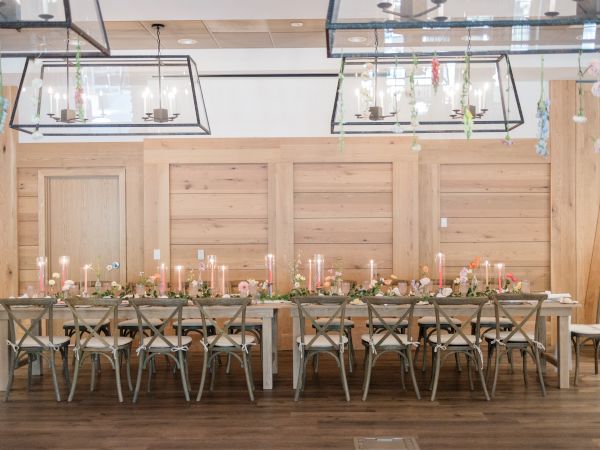 A decorated indoor event space with a long table set for guests, candles, flowers, greenery garlands, and lantern-style light fixtures.