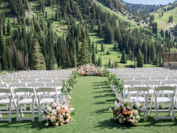 White wedding chairs line a sunny grass aisle with floral bouquets, set against steep green mountains and tall pine trees.