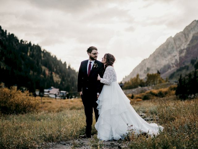 A bride and groom in wedding attire stand together outdoors in a mountain valley, gazing at each other.