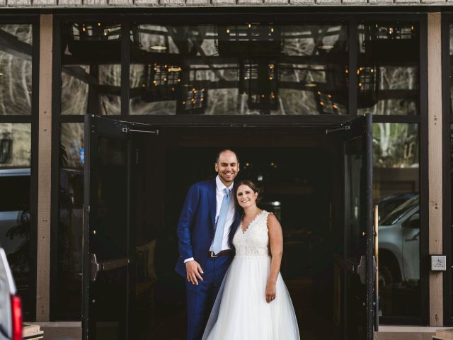 A smiling couple in wedding attire stands by open glass doors with warm lights behind them.