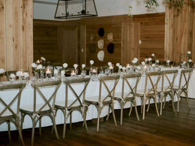 An elegant wedding reception setup with long banquet tables, white tablecloths, gold chairs, candles, and white flowers in a rustic hall.