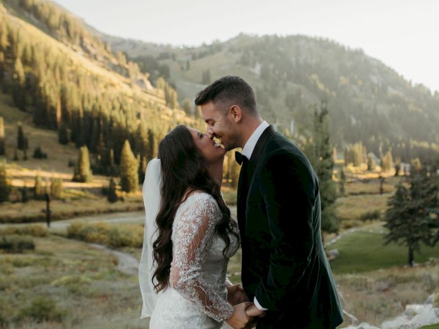 A bride and groom share an intimate kiss outdoors in a mountain valley, surrounded by trees and golden sunset light.