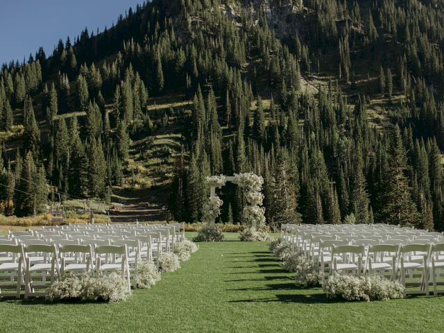An outdoor wedding ceremony setup with white chairs, floral archway, and a backdrop of tall alpine mountains and pine trees.
