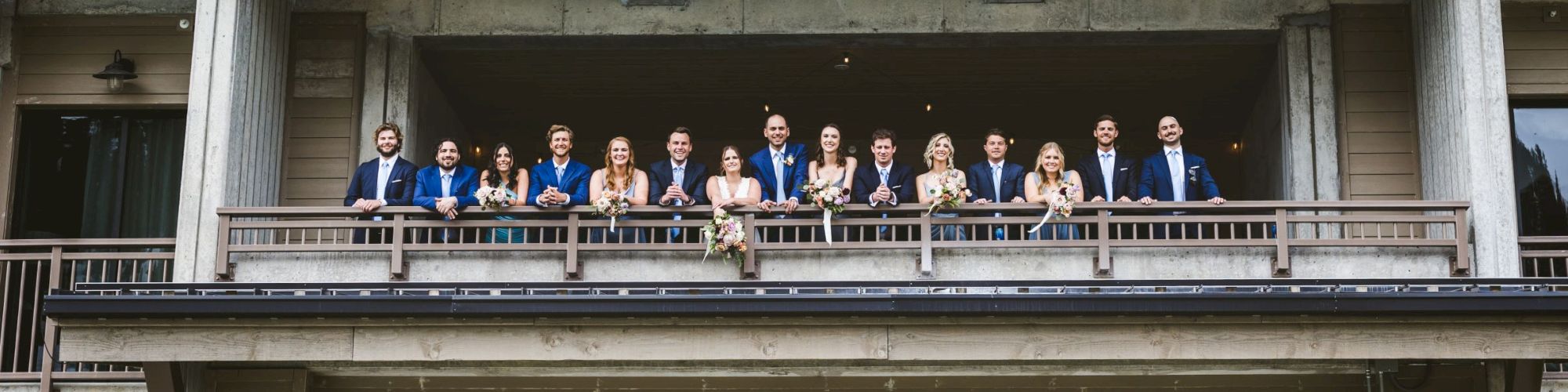 A wedding party in blue suits and white dresses stands on a building&rsquo;s balcony holding bouquets, smiling for a group photo.